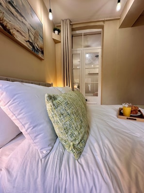 Ein Bett mit weißen Leinentüchern, ein grünes Kissen mit Struktur, ein Tablett mit Frühstück und ein Gemälde an der Wand.