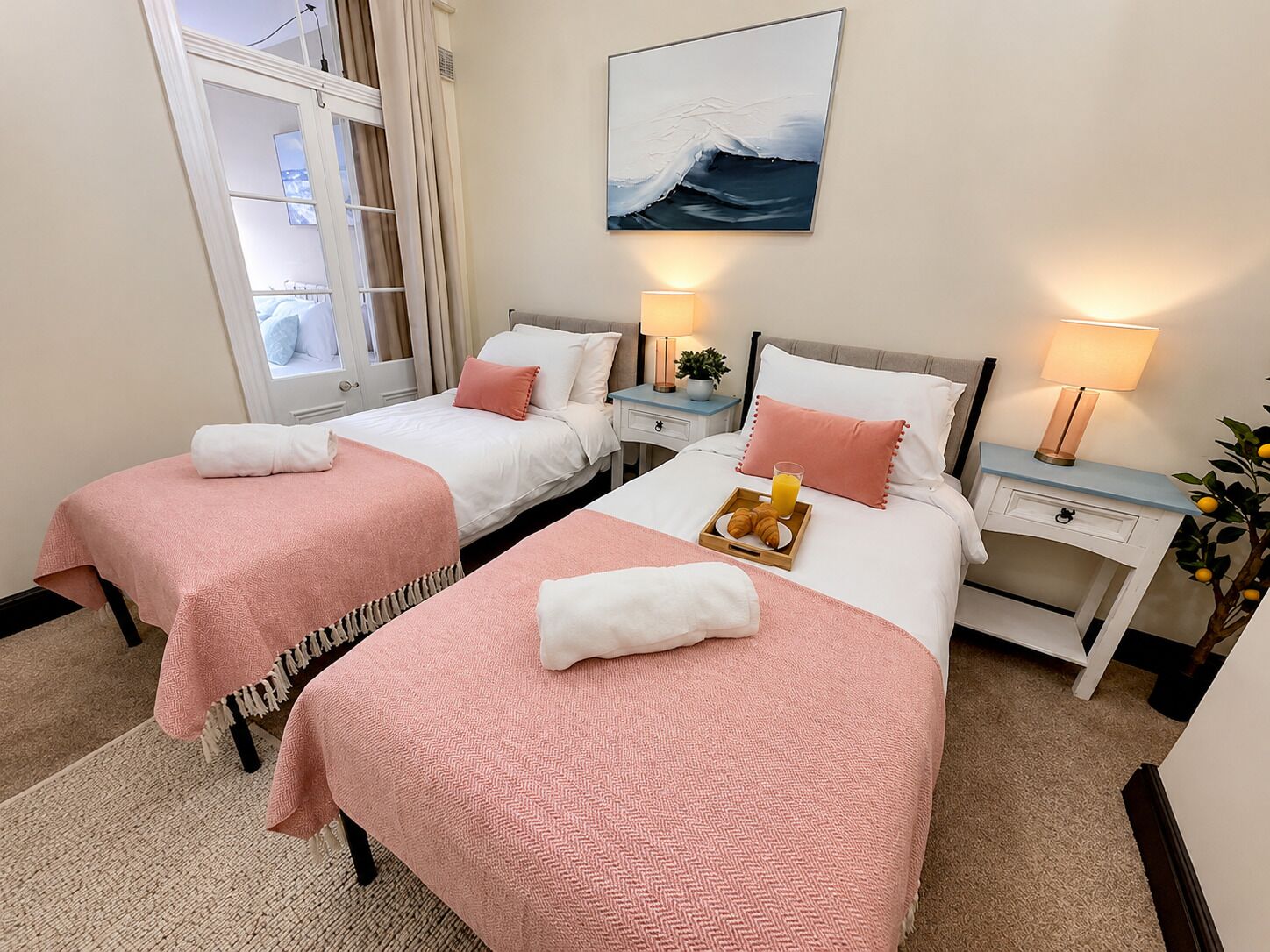 A hotel room with two beds, each with a pink blanket and white pillows, a bedside table with a lamp, a tray with croissants and juice, and a painting of a wave on the wall.