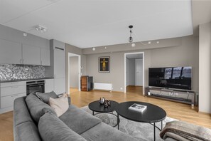 A modern living room with a grey sofa, two black coffee tables, a flat-screen TV, and a kitchen area with grey cabinets and a tiled backsplash. - The View - City center apartment with spectacular views (Tromsø)