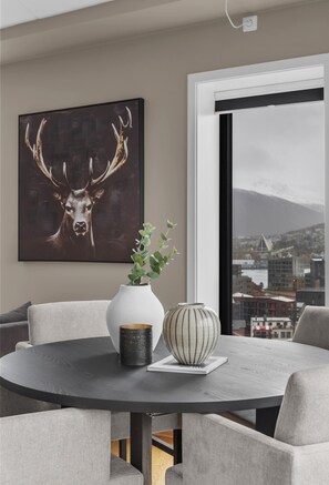 A modern dining area with a round table, two chairs, a vase with greenery, a decorative bowl, and a cityscape view through a large window. - The View - City center apartment with spectacular views (Tromsø)