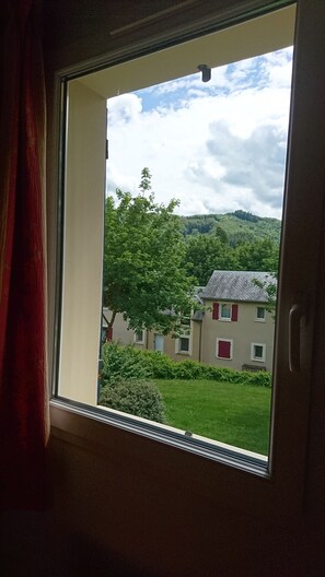 Ein Fenster mit Blick auf ein Gebäude mit roten Fensterläden, einer grünen Rasenfläche und eine Hügel im Hintergrund.
