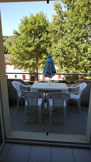 A balcony with a table and chairs, an umbrella, and a view of trees and buildings. - Apartment between Aubrac and the Lot Valley (Saint Geniez d'Olt et d'Aubrac)