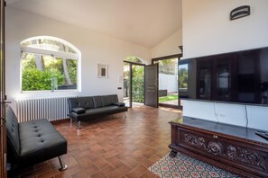 A spacious living room with a large TV, a sofa, and a window overlooking a garden. - Infinity Sea Views, Jacuzzi & Sunset Over Cefalù (Sicilia)
