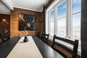 Ein Esszimmer mit einem langen Tisch, Stühlen, einer gewebten Tischmatte, einer Vase mit getrockneten Blumen, einem aufgehängten Hirschkopf und einem Fenster mit Blick auf Schnee.