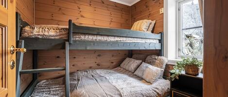 A bunk bed with a window view, wooden walls, and a plant on a dresser.