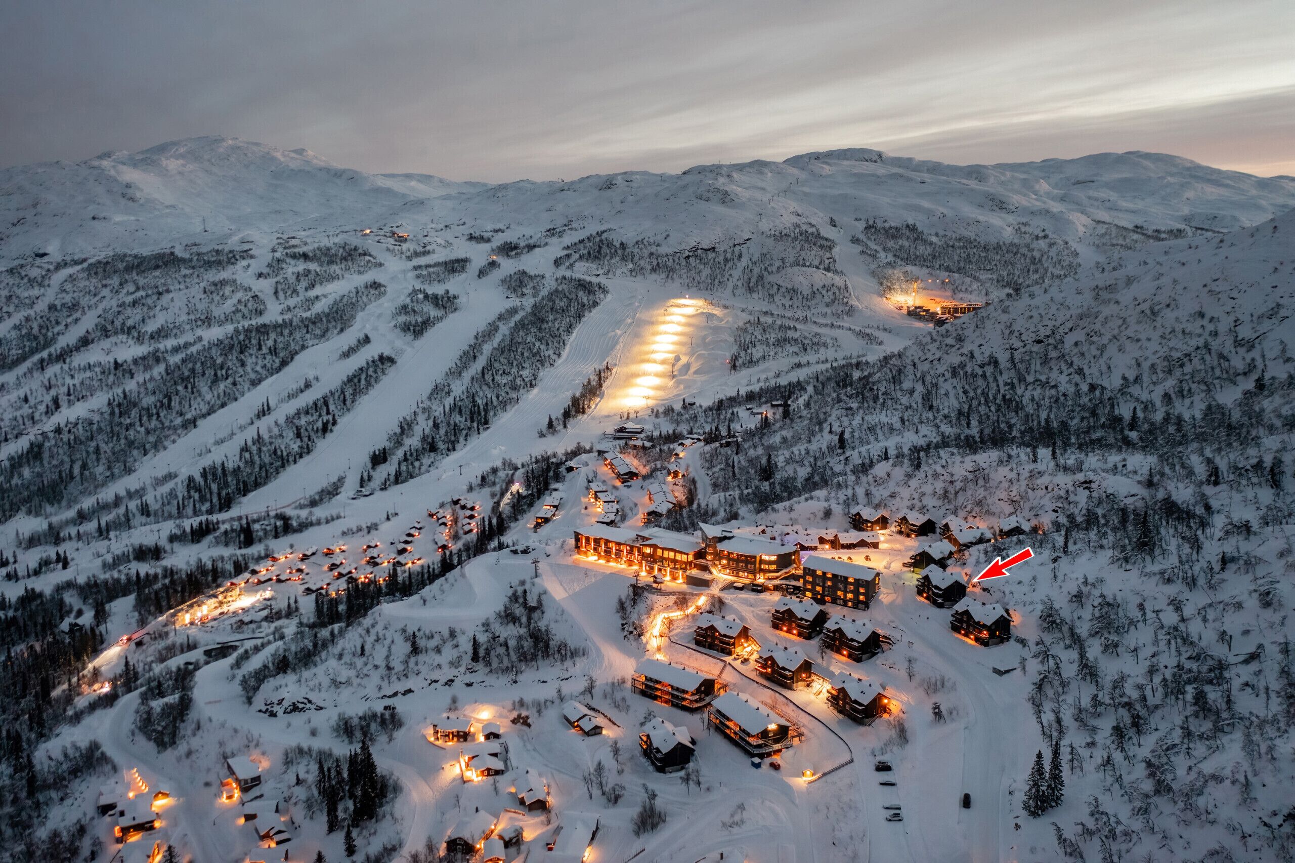 Ein schneebedecktes Bergresort mit mehreren Gebäuden, darunter eine große Lodge und kleinere Holzhütten, verbunden durch kurvenreiche Straßen.