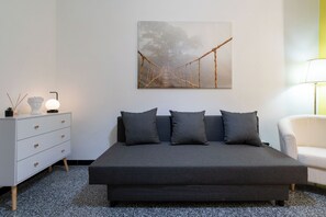 Une chambre moderne avec un canapé-lit gris, une commode blanche, une lampe et un tableau représentant un pont.