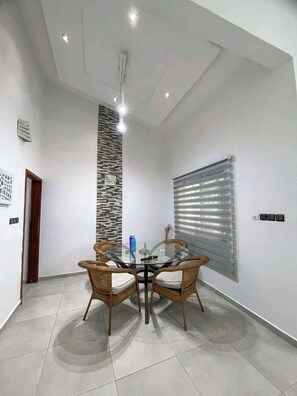 A modern dining area with a glass table surrounded by wicker chairs, a vertical textured wall, and a large window with blinds. - Votre havre de paix avec élégance et confort réunis (Abomey Calavi)