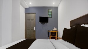 A hotel room with a bed, a television mounted on the wall, a small wooden table, and a door. - Deluxe With Balconyaps In (Prayagraj)