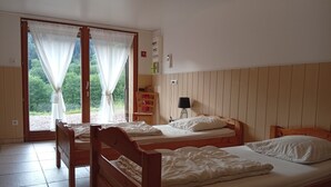 Ett rum med två sängar i trä, en stol, ett bord, en lampa och utsikt över grönskan genom ett fönster med tunna, genomskinliga gardiner.