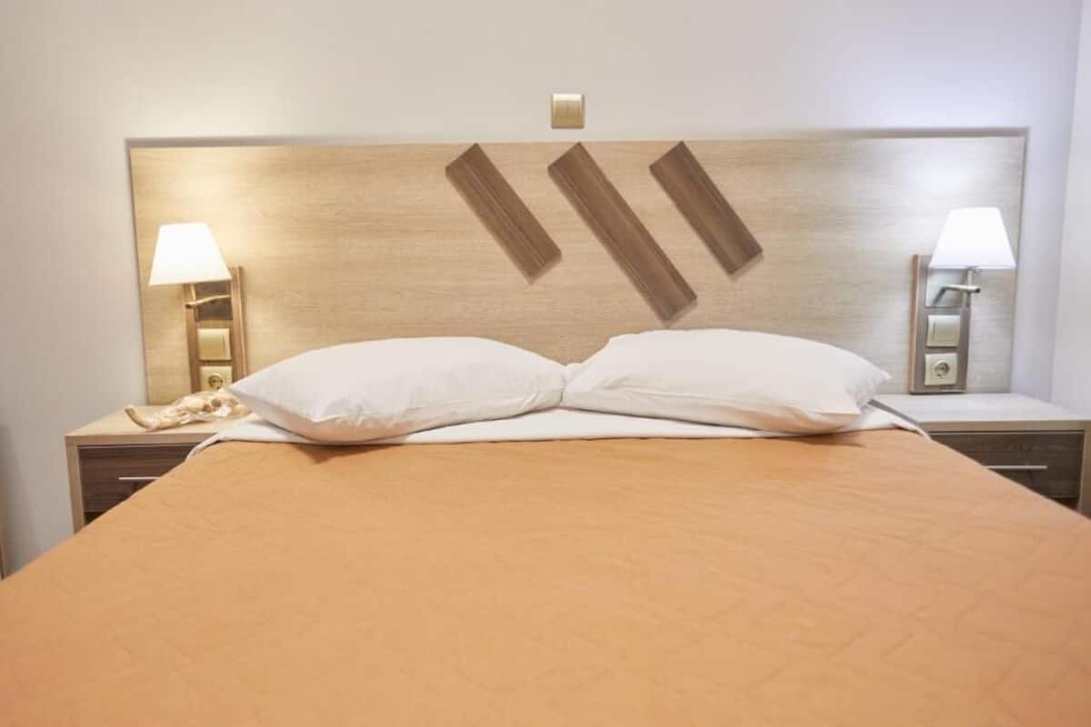 A hotel room with a large bed, two bedside lamps, a nightstand with a phone, and a wall-mounted light switch.