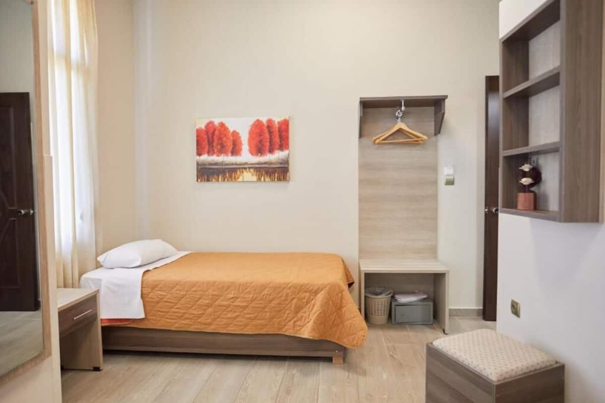 A bedroom with a bed, a hanging garment rack, a small bench, a wall-mounted shelf, and a painting.