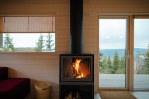 A wood-burning stove with a glass front, a red sofa, a woven basket, and a view of trees and mountains through a window. - Mountain cabin near Hafjell with view and hot tub (Øyer)