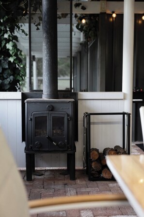 A wood-burning stove with a metal chimney, a stack of firewood, and a white wall with a window. - Cosy Winter Retreat  Fire, Tea & Sauna (preston beach)