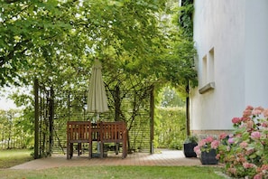 Eine Holzbank unter einer Pergola mit Sonnenschirm, umgeben von Grünpflanzen und einem weißen Gebäude.