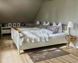 Ein Schlafzimmer mit schrägverdeckter Decke, einem Bett mit Holz-Kopfteil, Nachttischen, einer Lampe und einem Teppich.