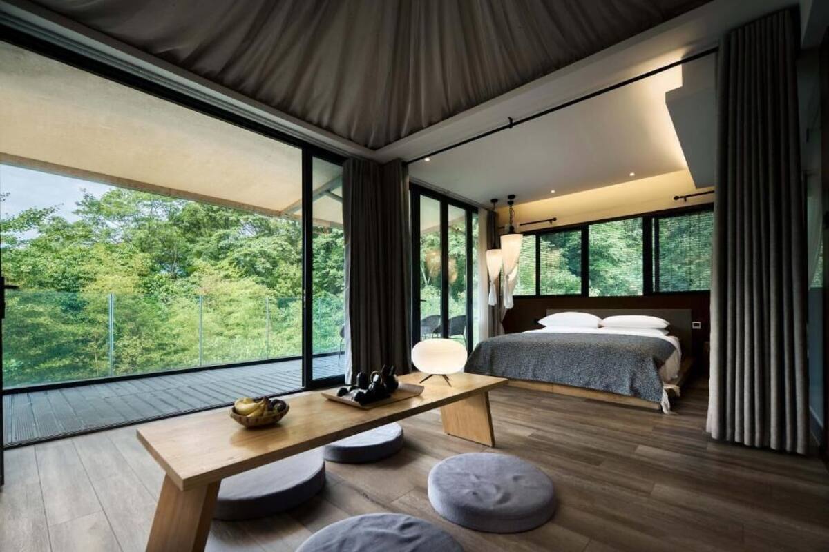 A modern bedroom with a large window, a bed, a table, and seating areas.