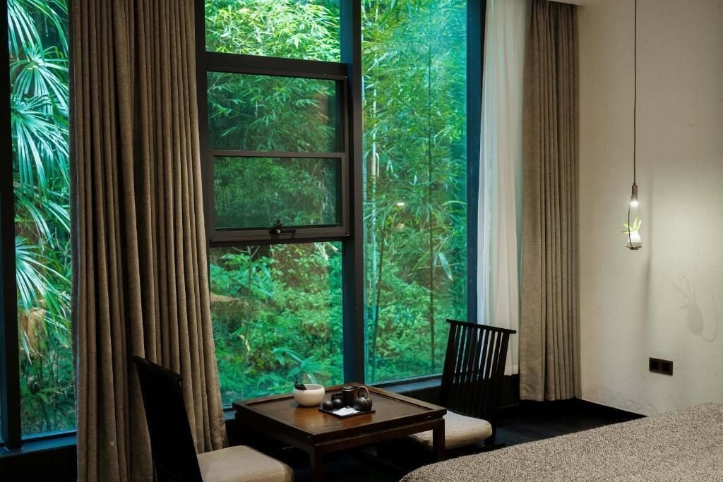 A room with a large window, a small table with a teapot, a chair, and a bed.