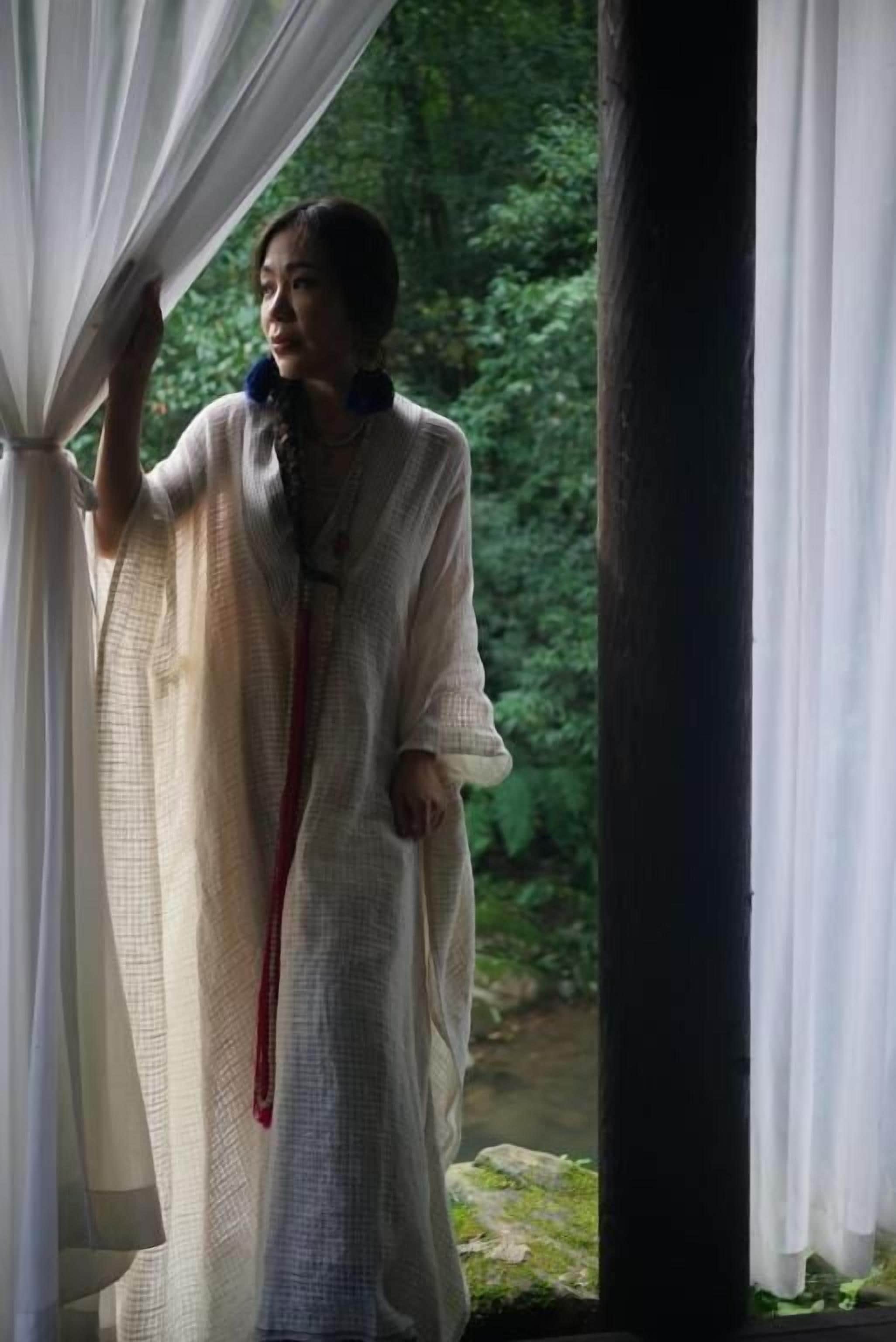 Una persona de pie junto a una ventana con cortinas, vista a un paisaje verde.