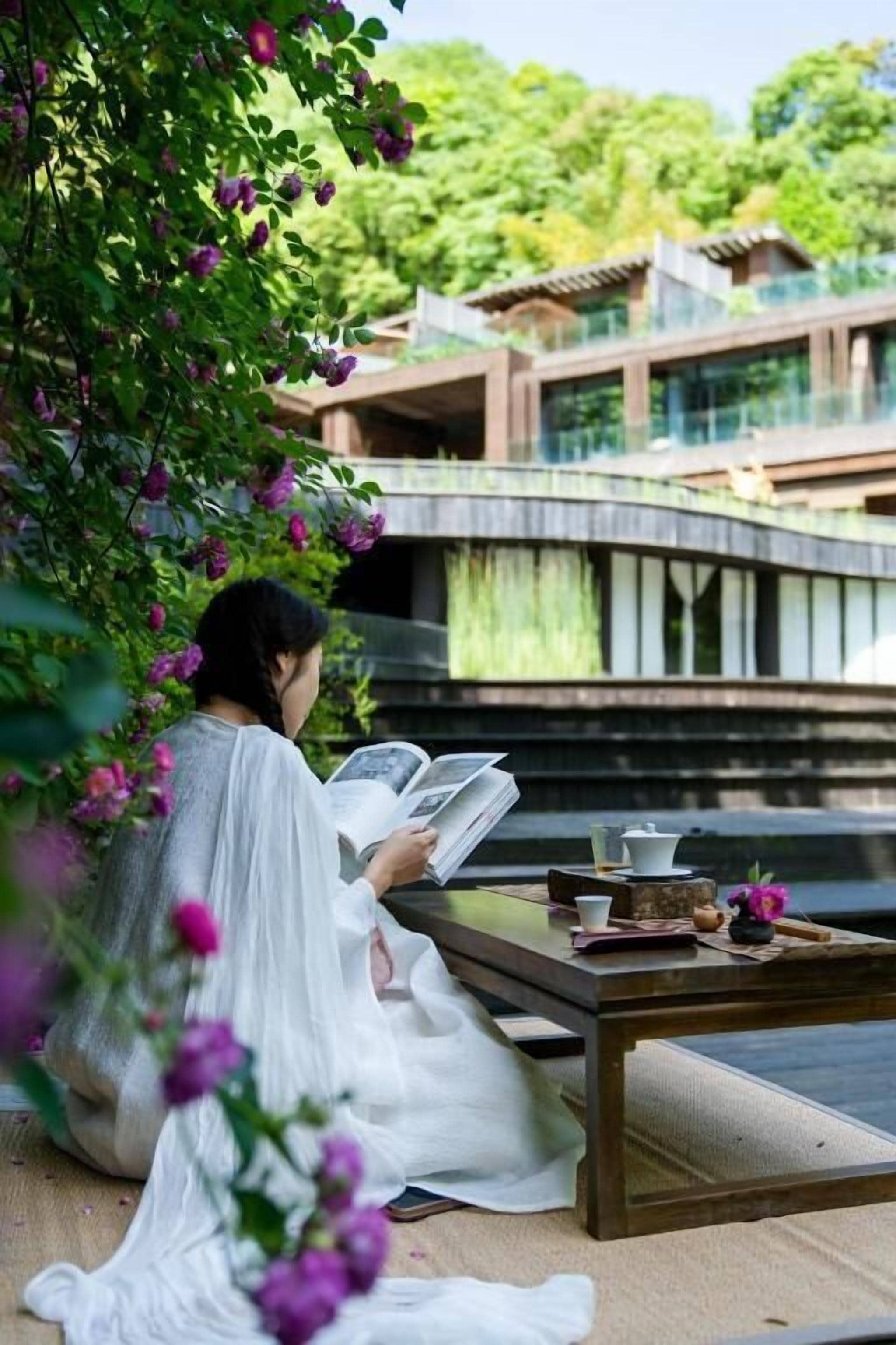 Una persona leyendo un libro en un entorno tranquilo al aire libre, con una mesa, una taza y una tetera.