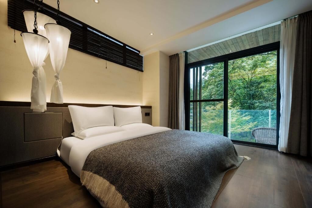 A modern hotel room with a large bed, a nightstand, a lamp, and a large window with a view of greenery.