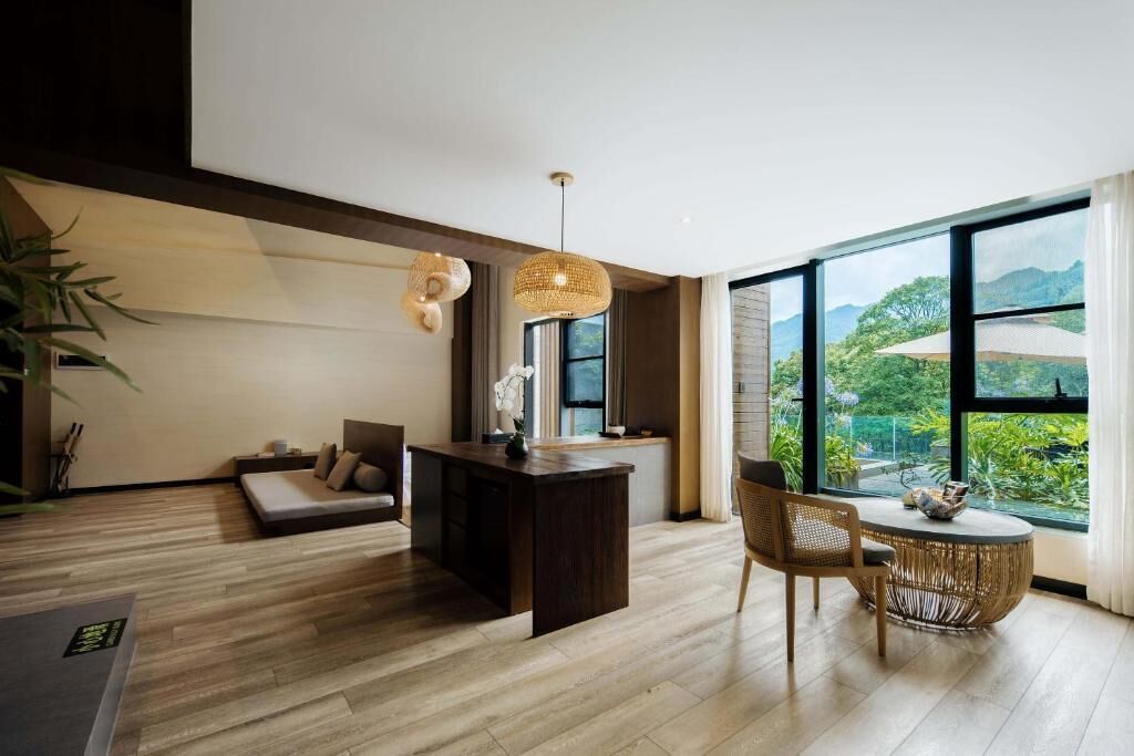 A modern living room with a wooden floor, a sofa, a dining table, and a large window offering a view of greenery and mountains.
