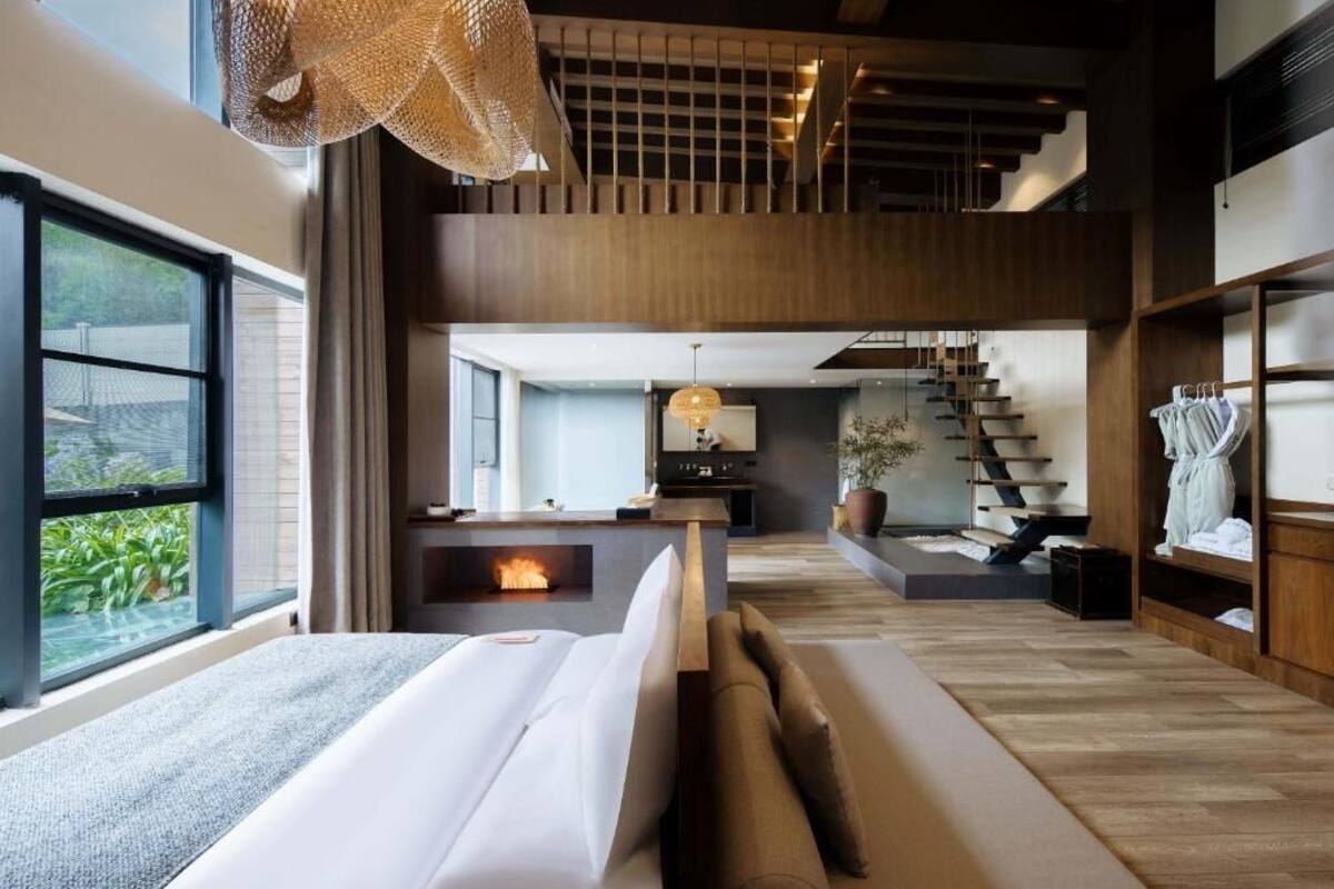 A modern hotel room with a large bed, a fireplace, a staircase, and a view of greenery.