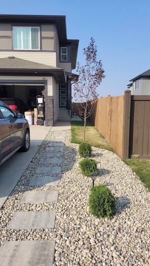A residential driveway with a car parked in the garage, a small tree, and a wooden fence. - Charming 1-bedroom Basement Suite in Spruce Grove, Alberta (Spruce Grove)
