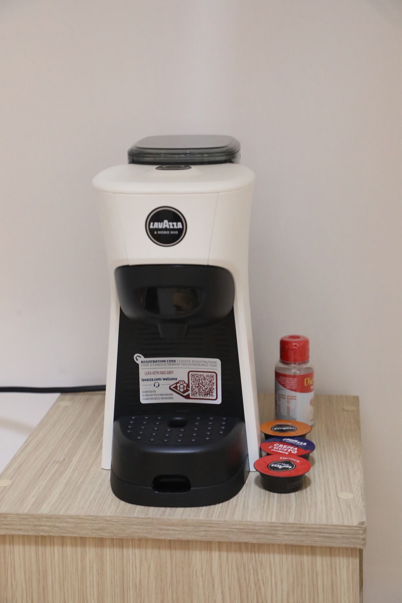 Une machine à café avec une étiquette indiquant un code d’enregistrement, un code QR et trois dosettes de café, le tout posé sur une surface en bois.