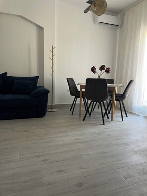 Een moderne woonkamer met een donkere bank, een eettafel met stoelen, een vaas met bloemen en een hoge staande lamp.