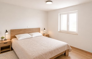 Ein Schlafzimmer mit einem hölzernen Kopfteil, zwei Nachttischen mit Lampen, einem großen Fenster mit Jalousien und einer gemusterten Bettdecke.