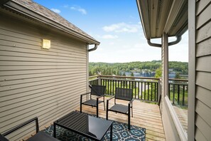 A balcony with a view of a lake and forest, wooden deck, and two chairs and a table.