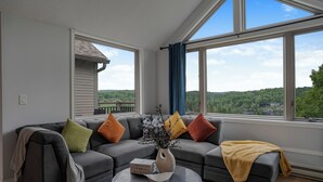 A modern living room with a gray sectional sofa, a coffee table, and a view of a green landscape through large windows. - Huntsville Lakeview Retreat | Jacuzzi & Games (Huntsville)