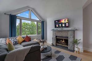 Una sala de estar con chimenea, un sofá con cojines de colores, un televisor de pantalla plana y un ventanal con cortinas.