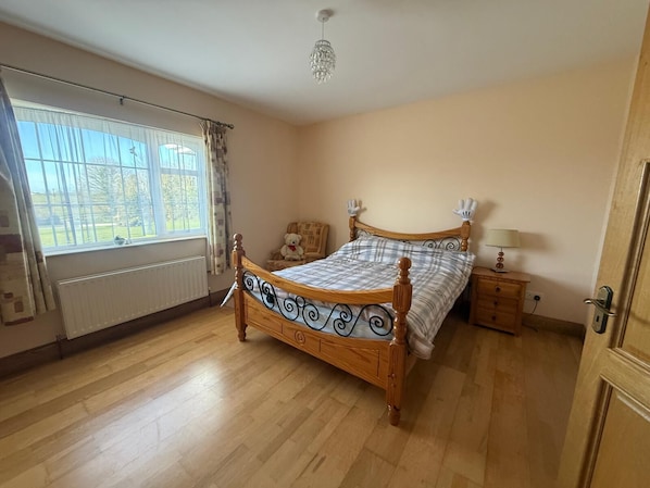 A bedroom with a wooden bed, a window with curtains, a radiator, a bedside table with a lamp, and a door.