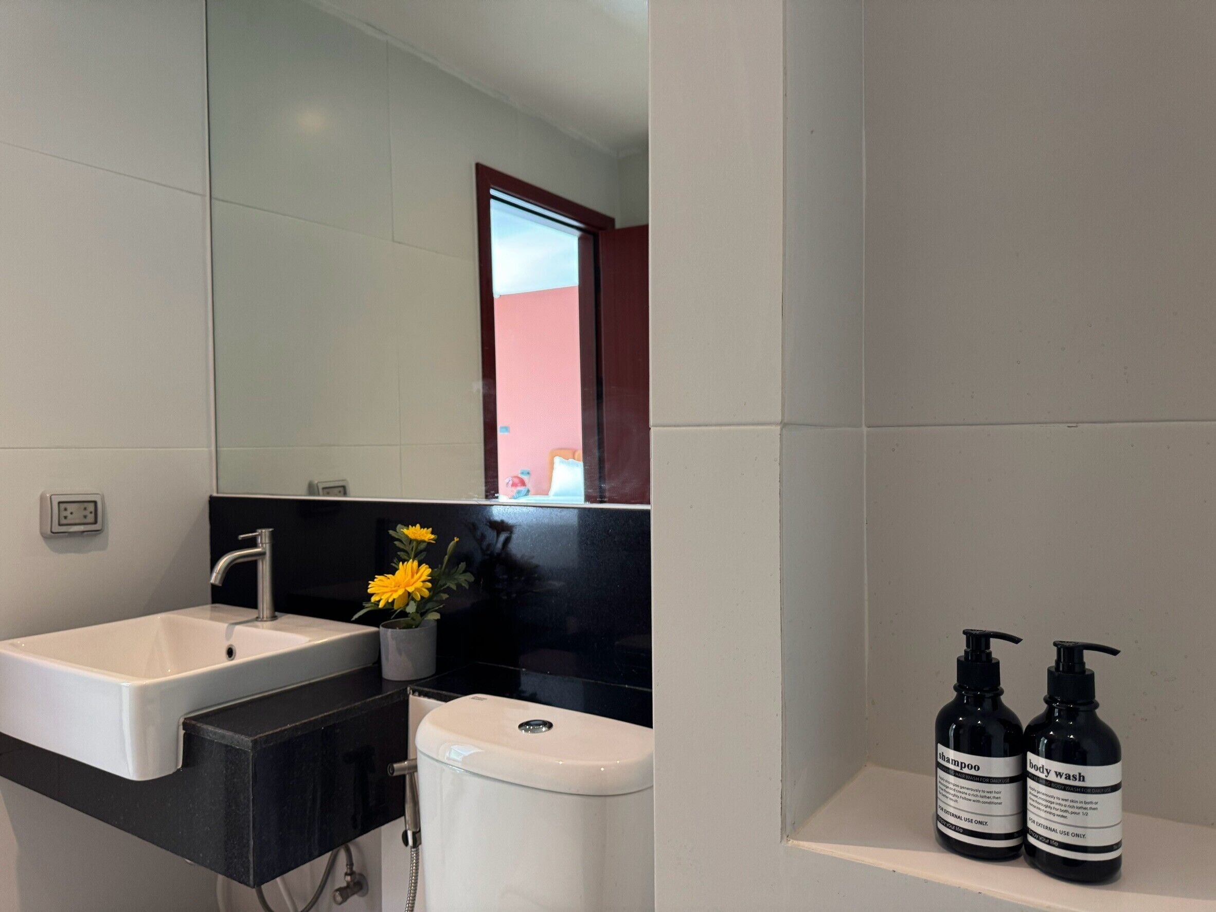 Un baño moderno con un lavamanos blanco, encimera negra y un inodoro. Se ven dos botellas de champú y crema corporal a la derecha.
