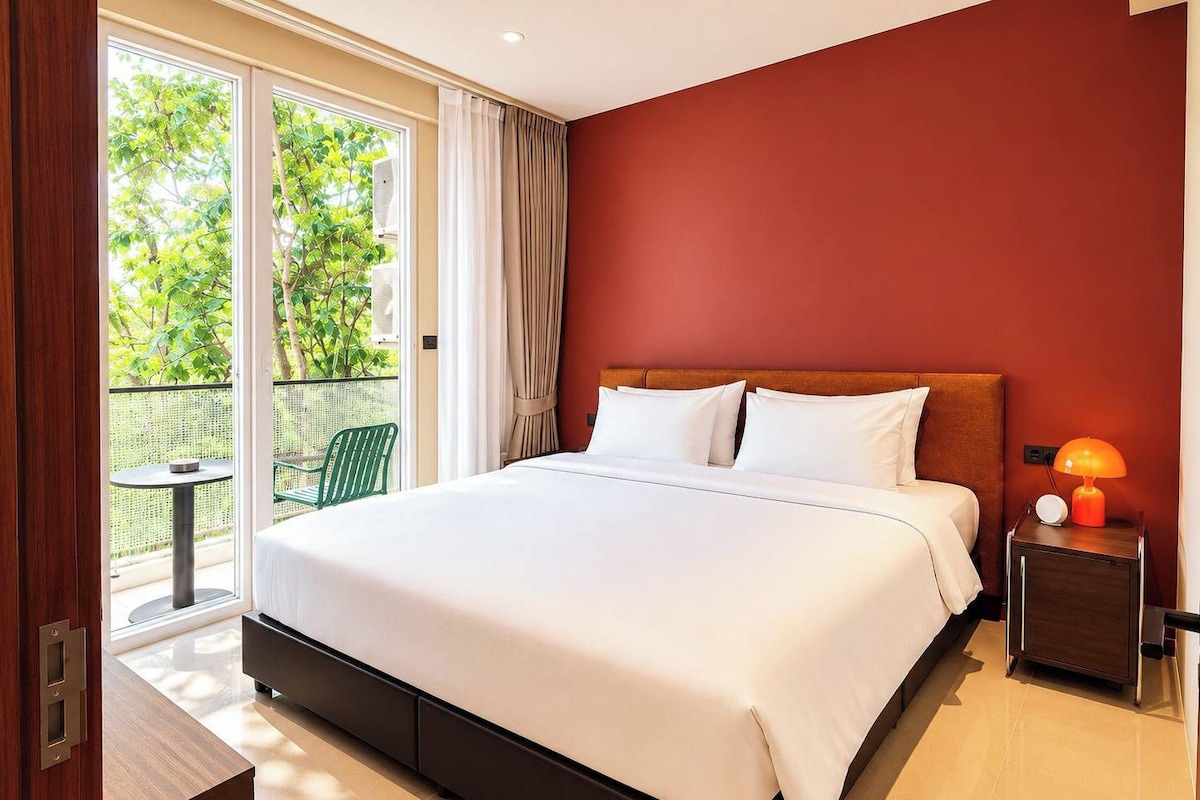 Habitación de hotel con una cama grande, una mesita de noche con lámpara, vistas a zonas verdes y un balcón con una silla.