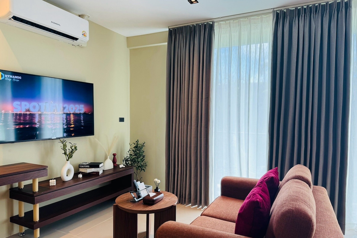 Una sala moderna con televisor de pantalla plana, un sofá, una mesa de centro y un amplio ventanal con cortinas.
