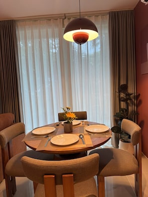 A dining area with a round table set for four, surrounded by chairs, with a pendant light above and a plant to the side. - Syama Mansion Kamala (Kamala)
