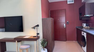 Un espacio habitable pequeño con kitchenette, un televisor montado en la pared, un escritorio con silla, una planta en maceta y una puerta.