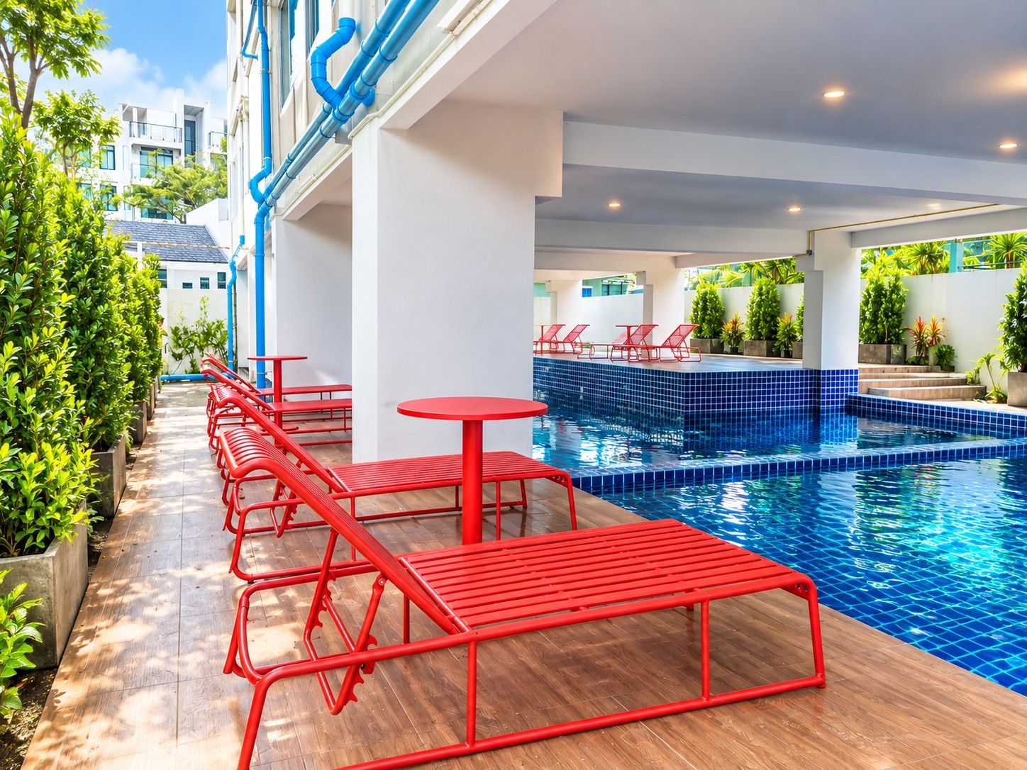 Zona de piscina con sillones rojas y mesas, rodeada de follaje y una piscina con baldosas.