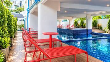 Zona de piscina con sillones rojas y mesas, rodeada de follaje y una piscina con baldosas.
