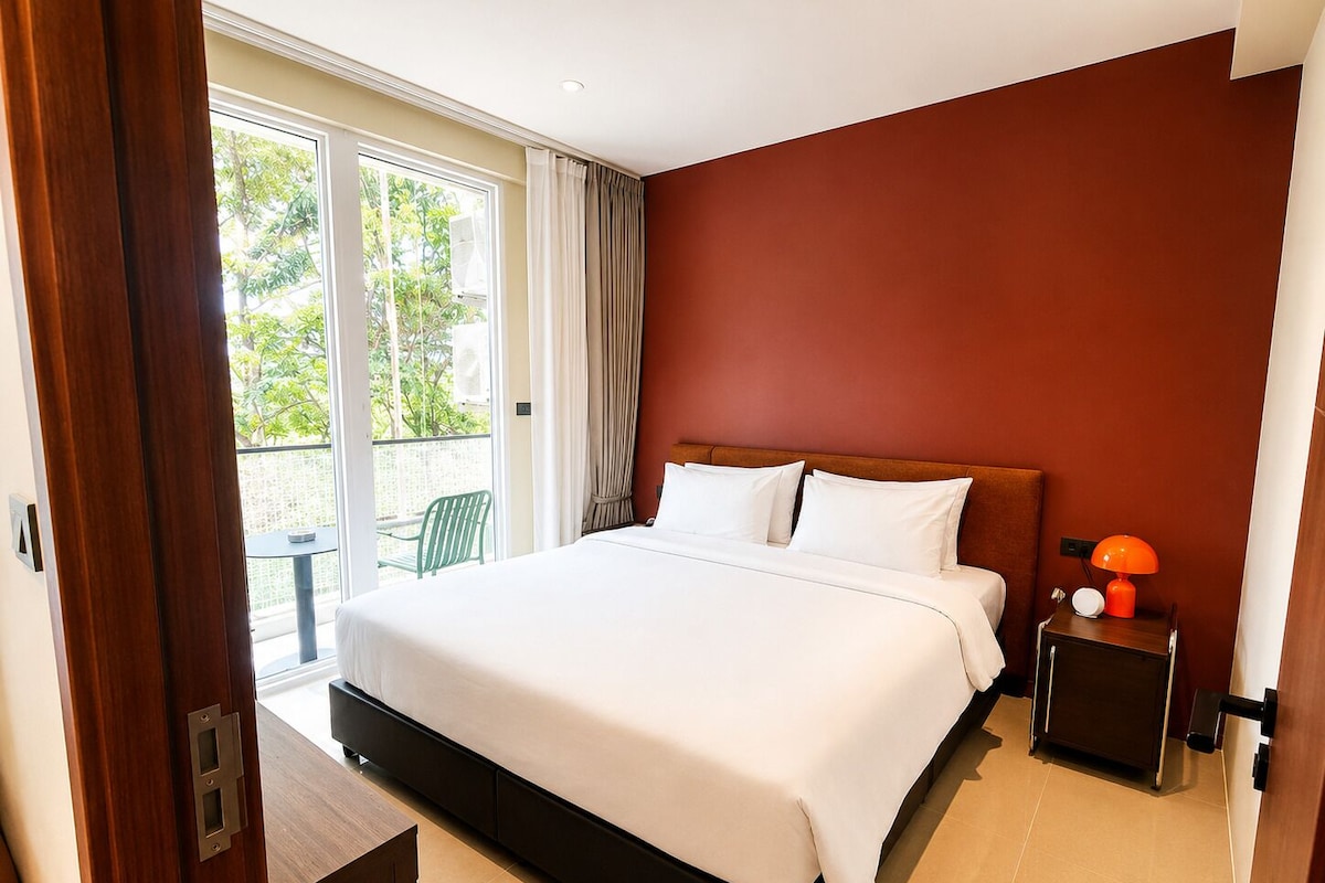 A hotel room with a bed, a nightstand, a lamp, and a view of greenery outside.