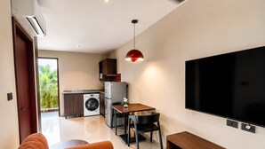 Un espacio habitable moderno con un sofá, un comedor con sillas, una lavadora, un refrigerador y un televisor de pantalla plana grande montado en la pared.