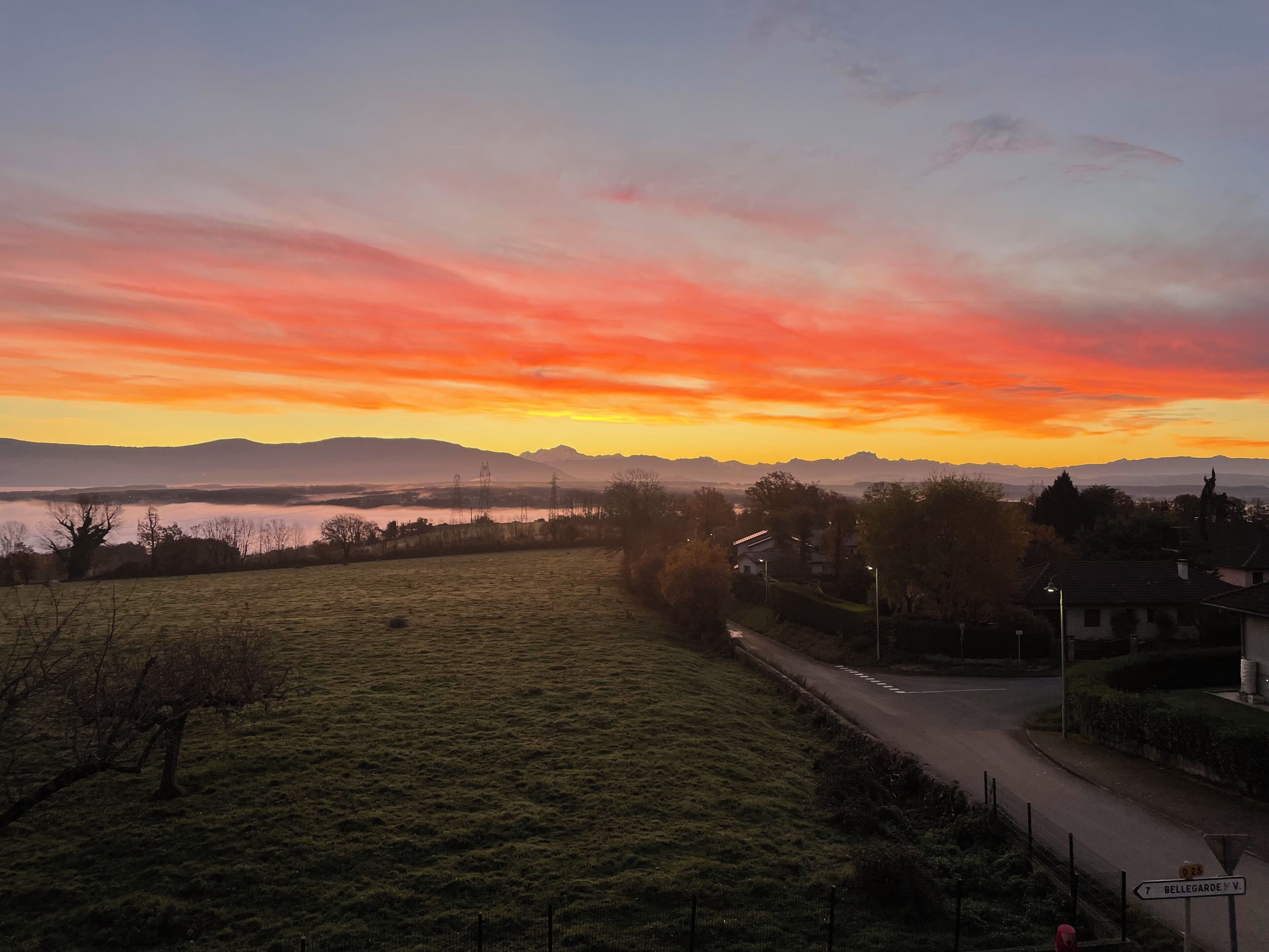 Ländliche Landschaft mit einer Straße, Häusern und einem Sonnenuntergang über Bergen.