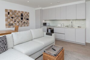 Ein modernes Wohnzimmer mit Sofa, Couchtisch und Küchenzeile mit weißen Schränken und Geräten.