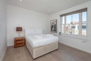 Ein Schlafzimmer mit Bett, Nachttisch, Lampe, einem gerahmten Bild und einem Fenster mit Blick auf Gebäude.