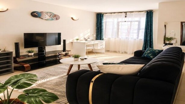A living room with a black sofa, a TV, a desk, and a plant. - Soothing 1-bedroom apartment in vibrant Nairobi perfect for relaxing stay (Nairobi)