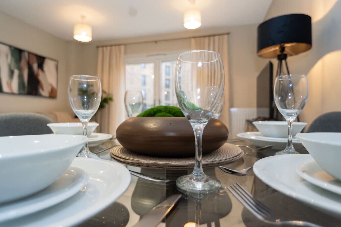 Een eettafel gedekt met witte borden, kommen en wijnglazen, met een decoratieve houten schaal en een groene plant als middelpunt.