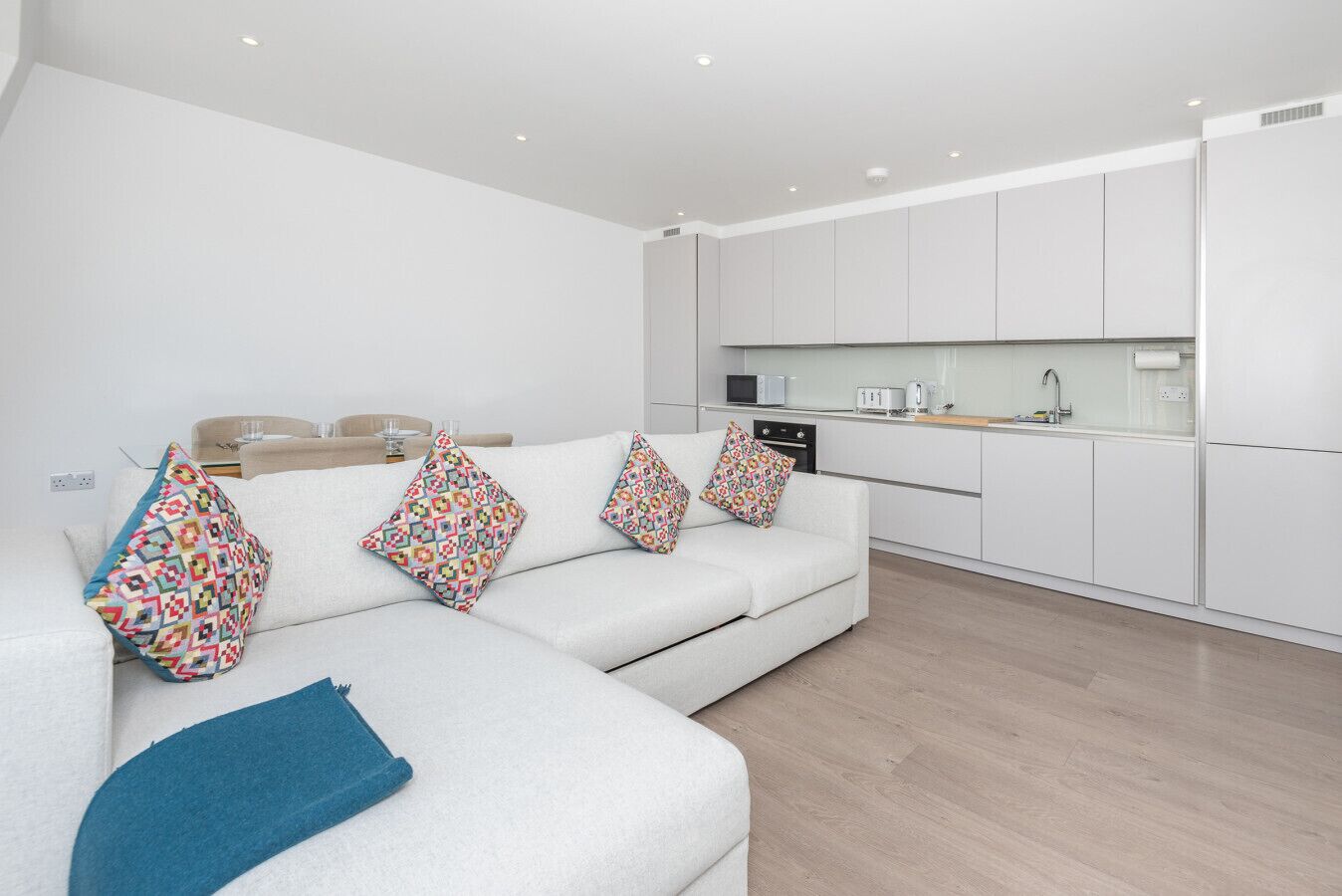 A modern living room with a white sofa, colorful pillows, a blue throw, and a kitchen area with white cabinets and appliances.